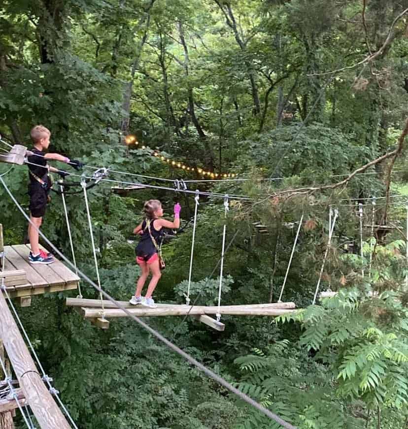 Two kids climbing across a rope course through the trees.