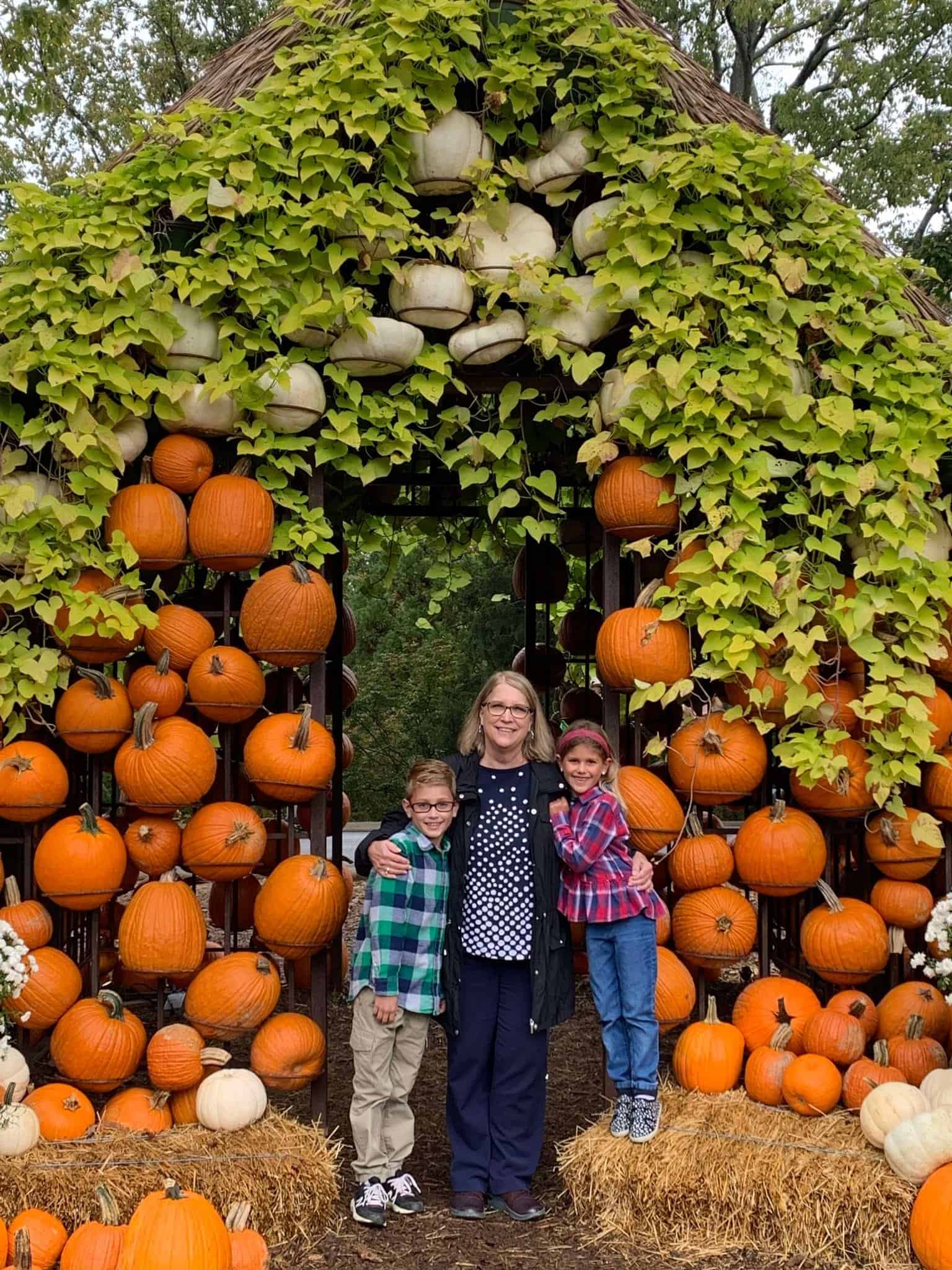 Grandma with two grandchildren standing in a house that is made out of dozens of pumpkins and wire.