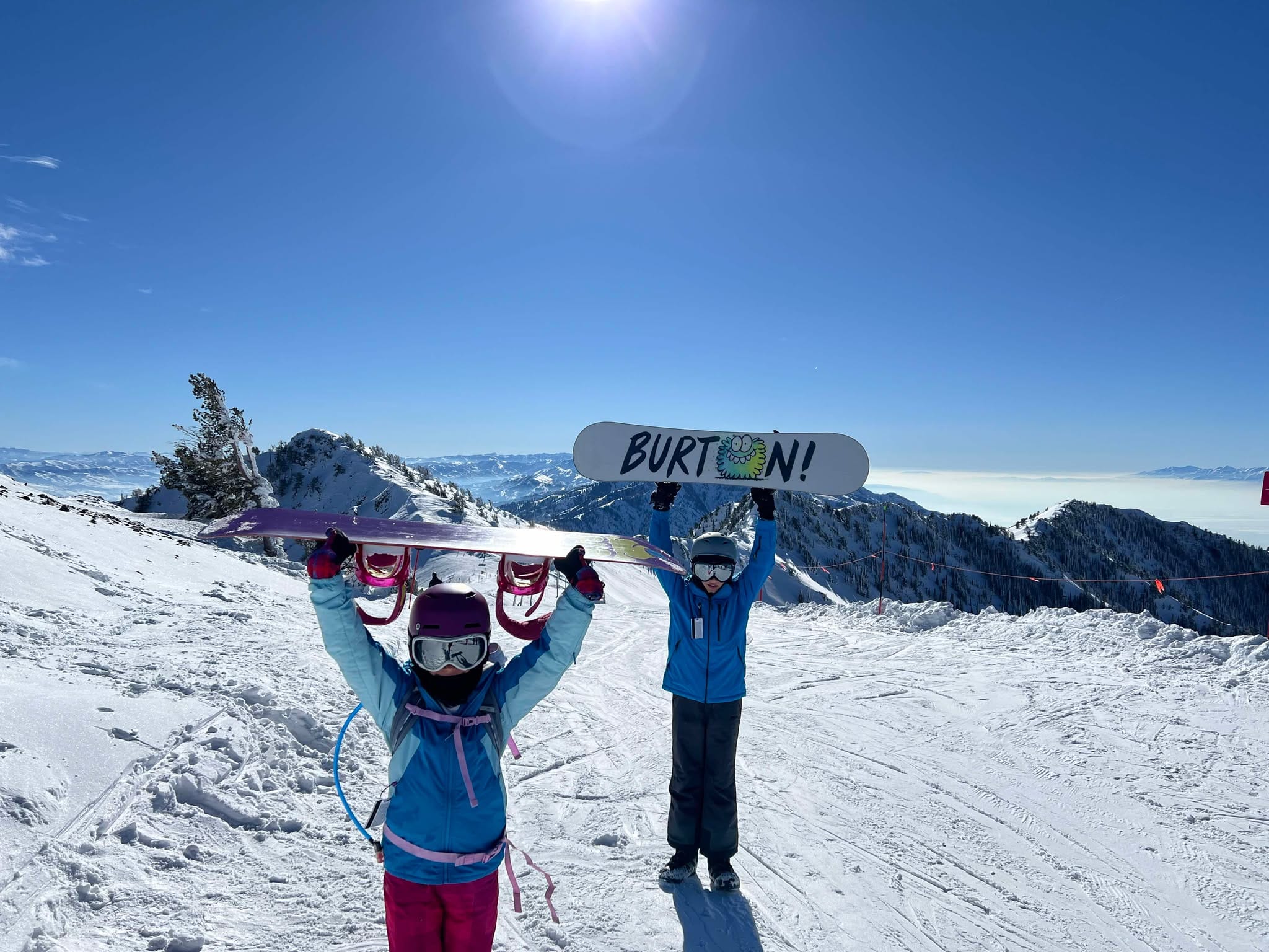 A 9 year old girl and a 12 year old boy holding up their snowboards at the top of Strawberry Run for their family snowboarding trip to Snowbasin Ski Resort.