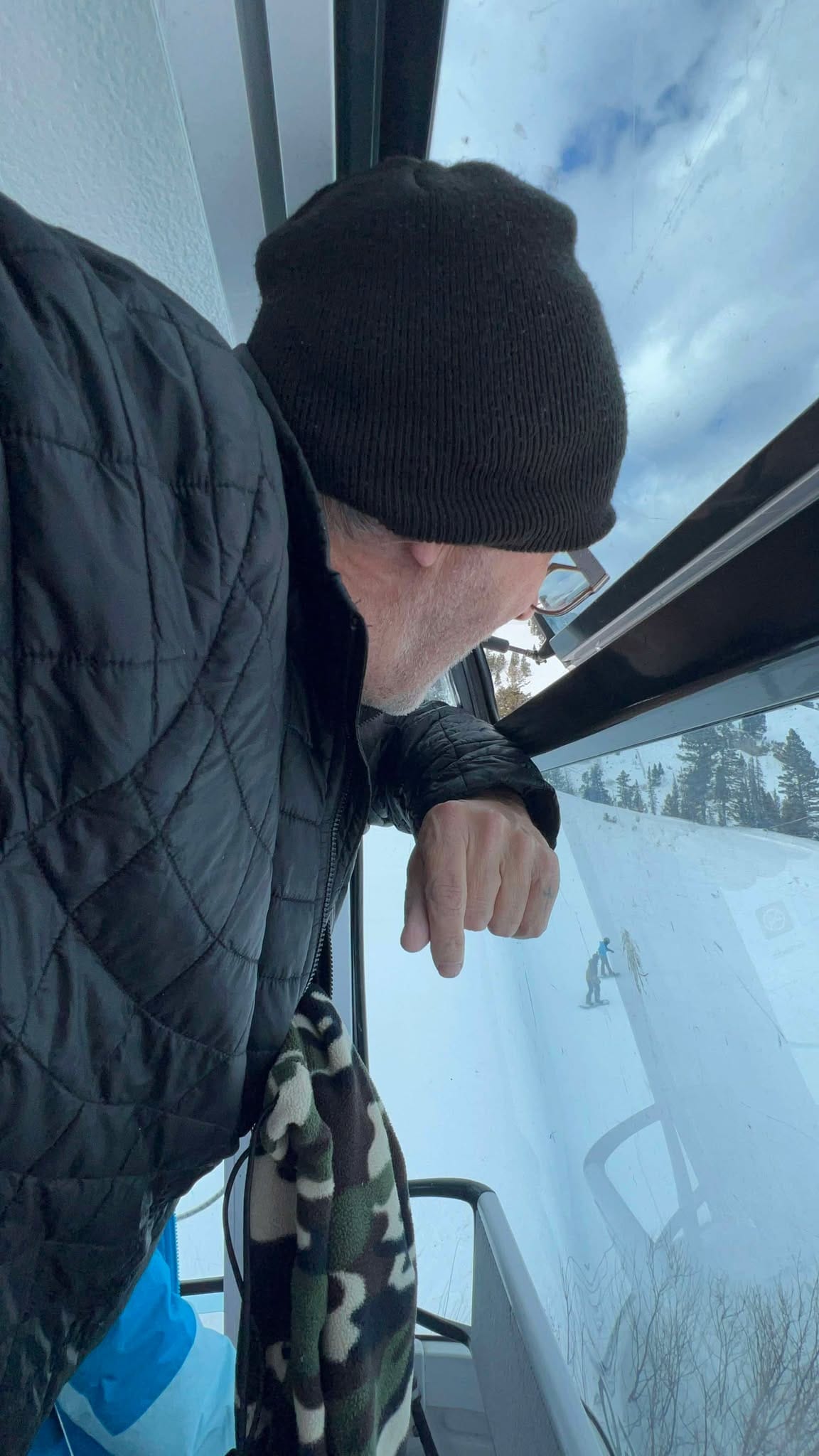 A grandpa on the gondola watches from above while his grandson snowboards below him.  
