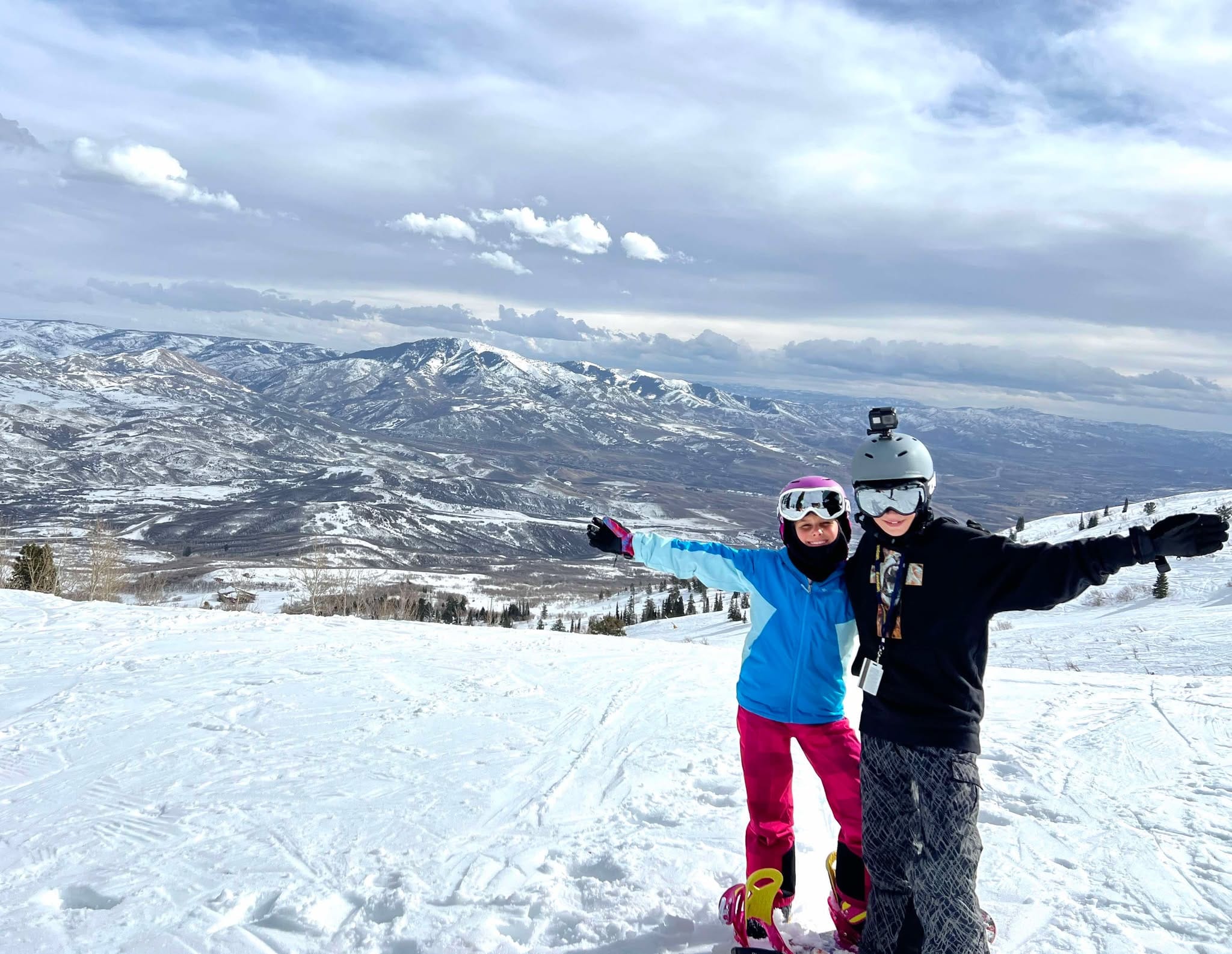 A boy and a girl holding out their arms at the top of the mountain during their family snowboarding trip to Snowbasin Ski Resort.