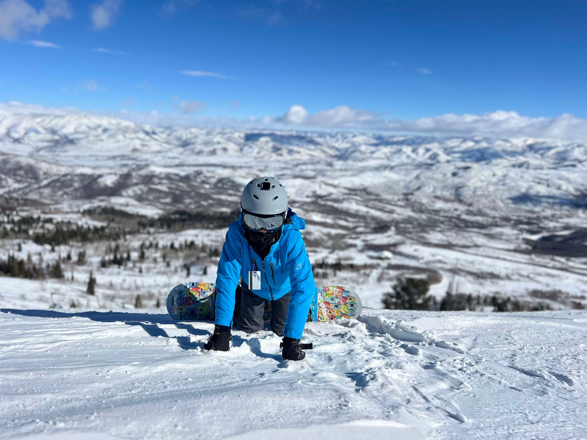 A boy on his knees looking at the camera at the top of the mountain while on his family snowboarding trip to Snowbasin Ski Resort.