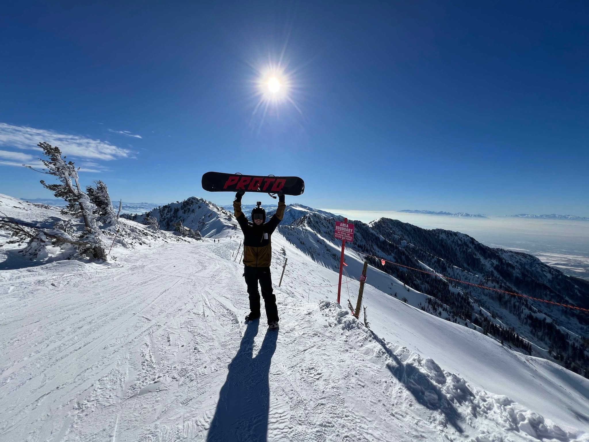 A man holding up his snowboard at the top of Snowbasin Ski Resort mountain while on a family snowboarding trip.