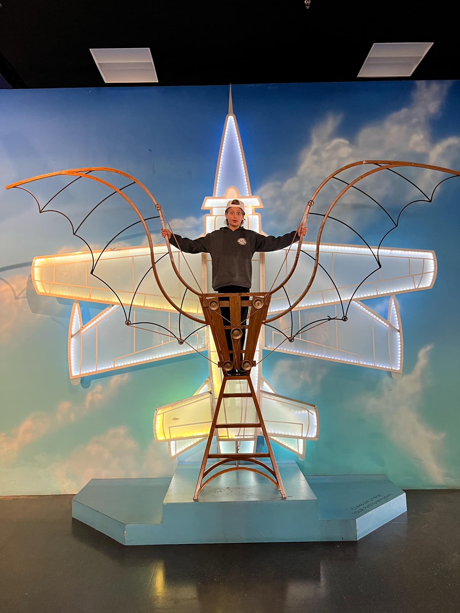 A boy standing in a Leonardo Da Vinci airplane contraption. 