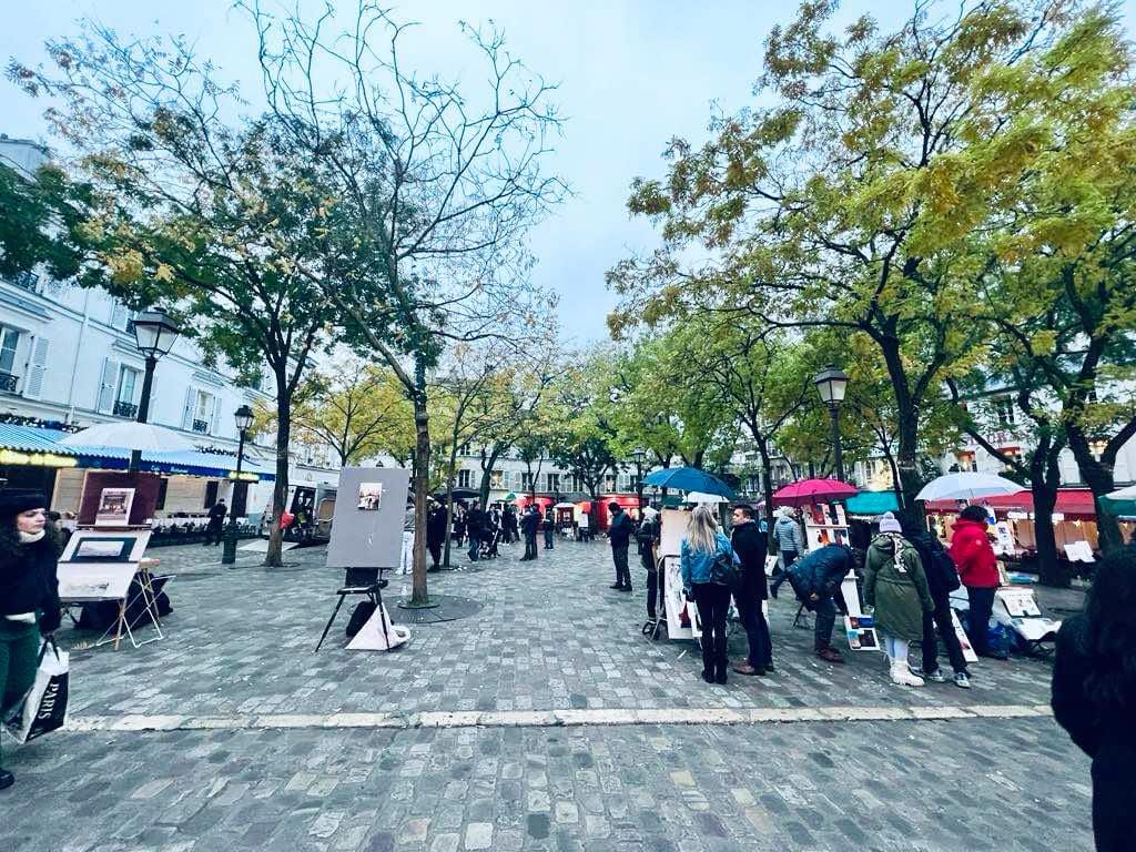 Picture of the square of Montmartre filled with cafes and live-action artists. This spot is free to enjoy and lots of people are doing that in the photo.