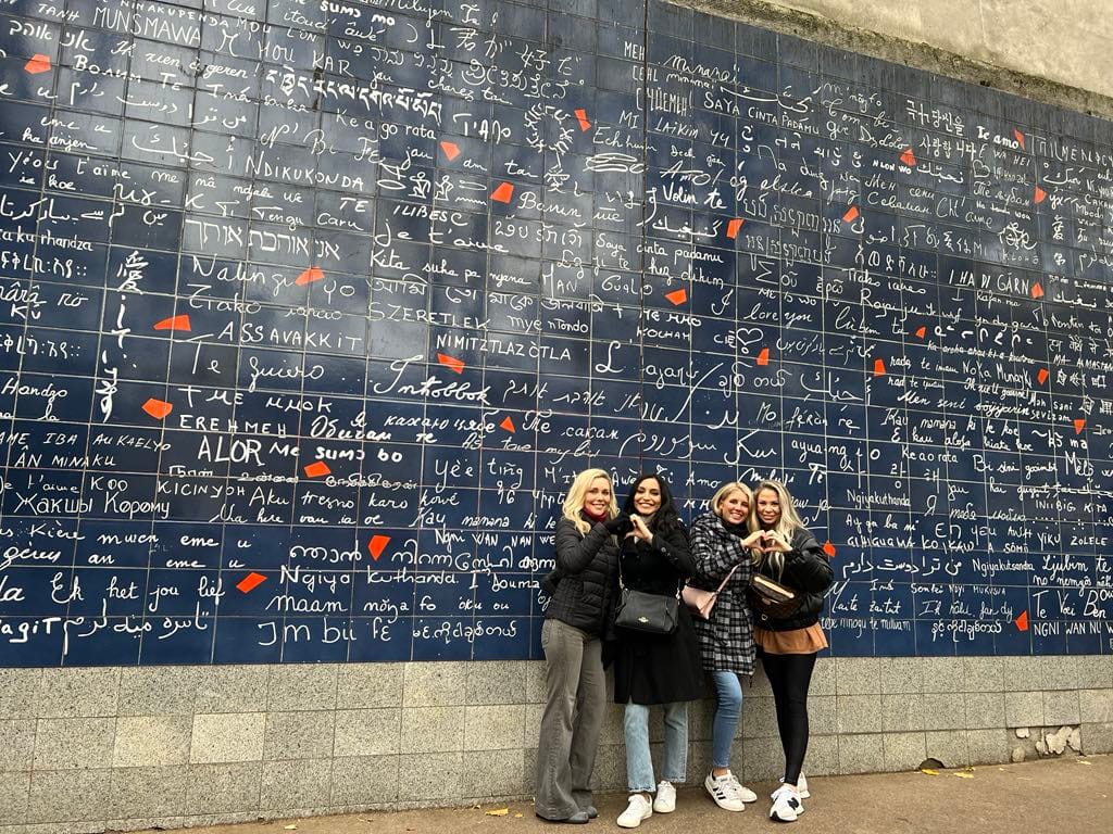 Four friends posing with heart hands at the "I love you wall" in Paris.