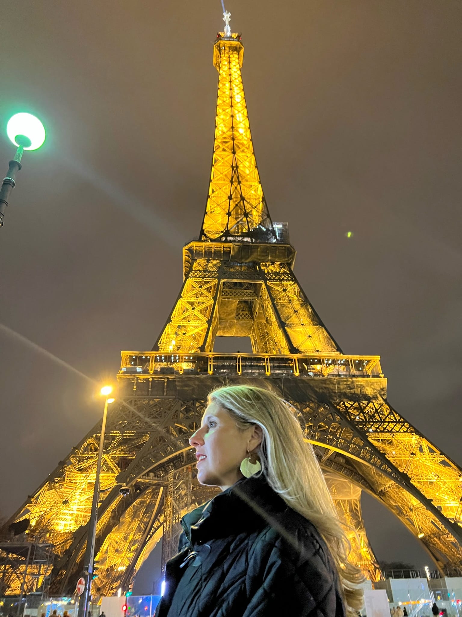 A very golden photo as the Eiffel Tower is lit up against a black/orangish sky. A girl with blonde hair stands in front of it looking off to the left side.