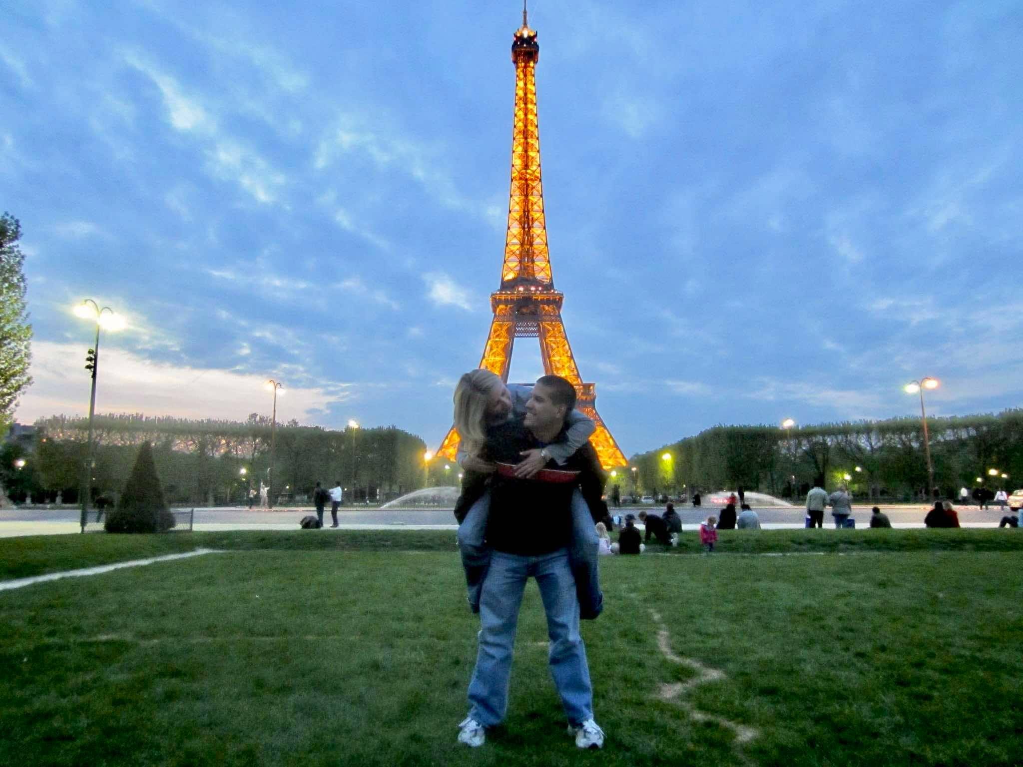 Girl on guy's shoulders in front of the Eiffel Tower lit up at night in Paris.
