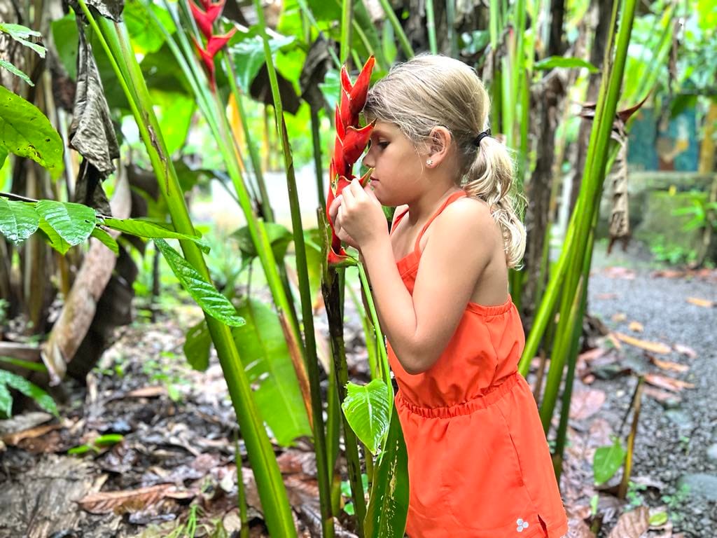 A little girl in a bright orange jumpsuit smelling a bright orange flower with plants all around her at the chocolate farm in Costa Rica.