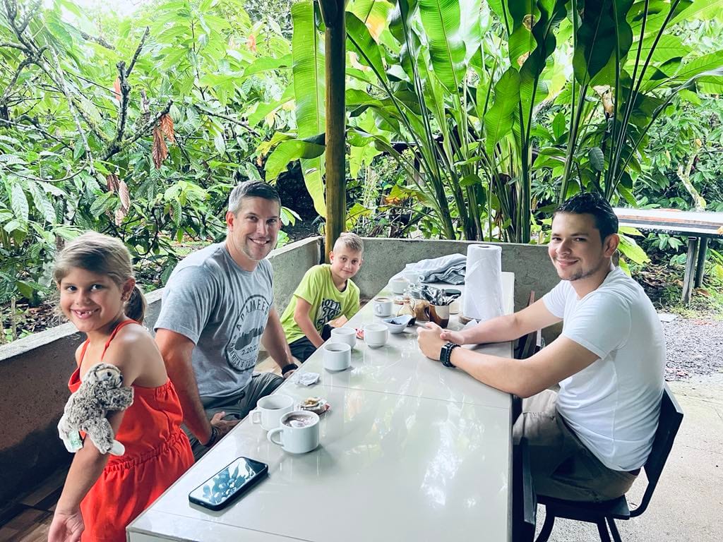 A dad and two kids with the tour guide sitting down at a table and enjoying chocolates and coffee at the chocolate farm tour. The little girl has a sloth stuffed animal on her arm.