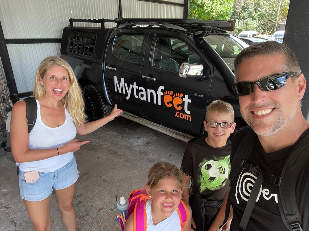A family of 4 standing in front of a car that says "my tan feet" while on their costa rica with kids trip.