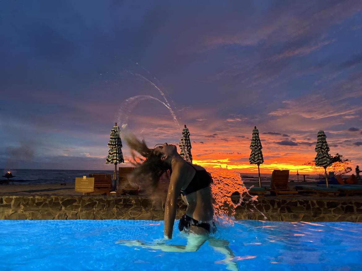 A girl in the pool flipping her hair back with an orange sunset behind her.