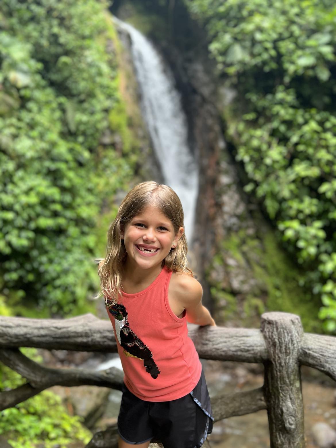 Little girl in front of a waterfall with a bright orange shirt that has a toucan on it.