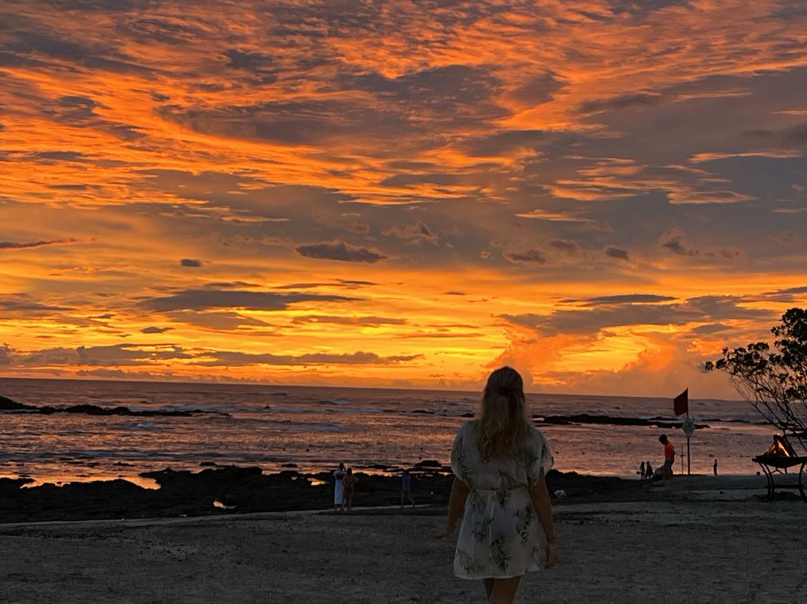 A mom walking on the beach with the orange sunset ahead of her.