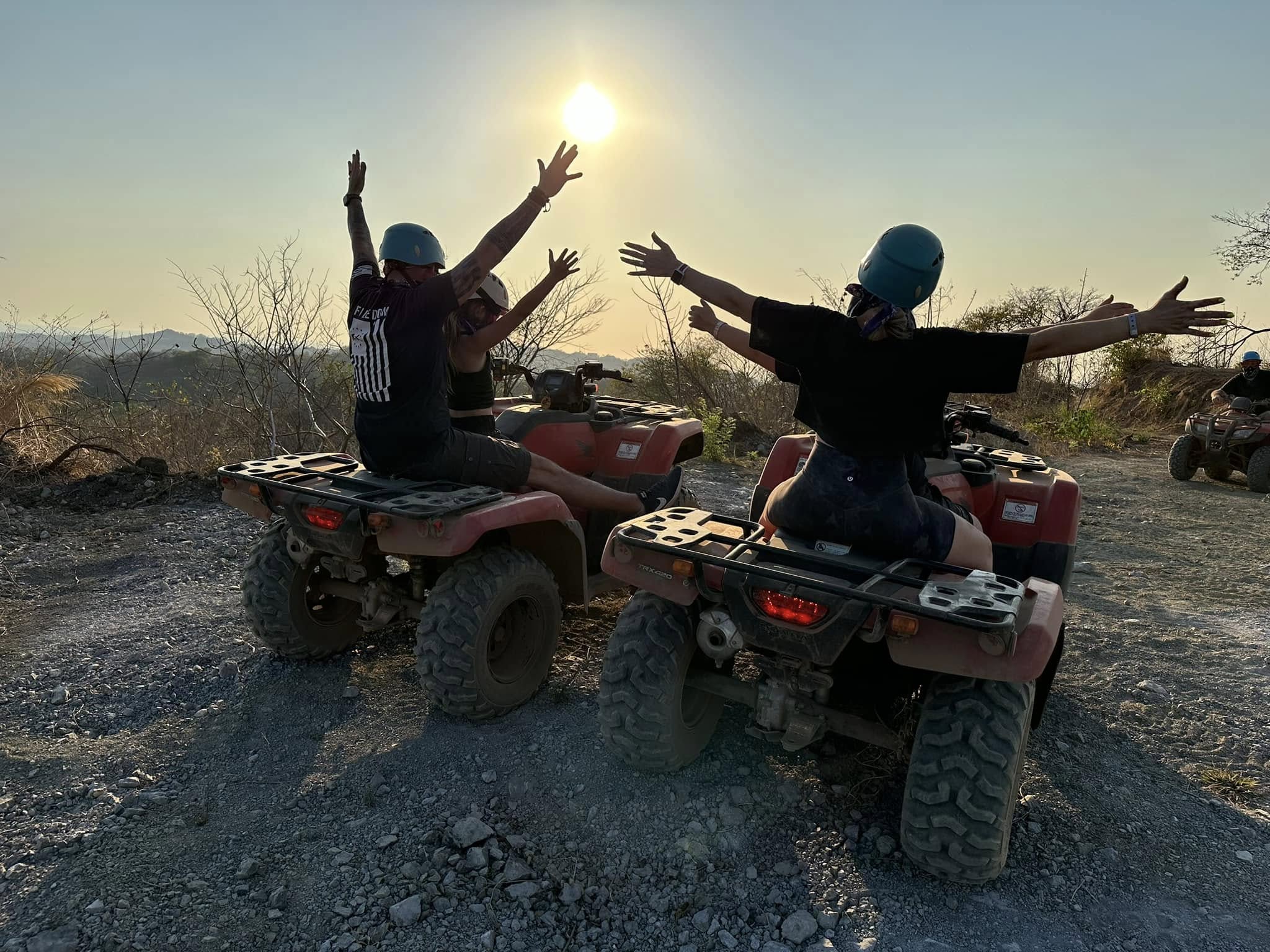 A family of 4 on 4-wheelers with the sunset ahead of them. They have their arms stretched out for fun.