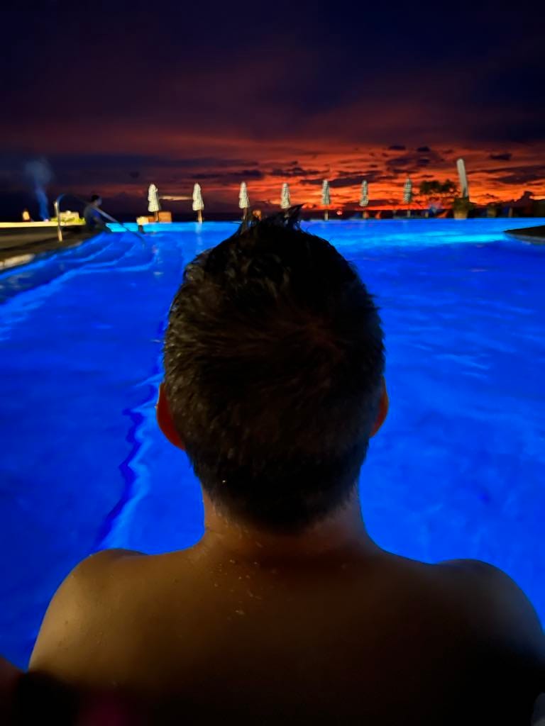 A view of a man's head with the blue pool in front of him and beyond that a bright orange sunset on the ocean.
