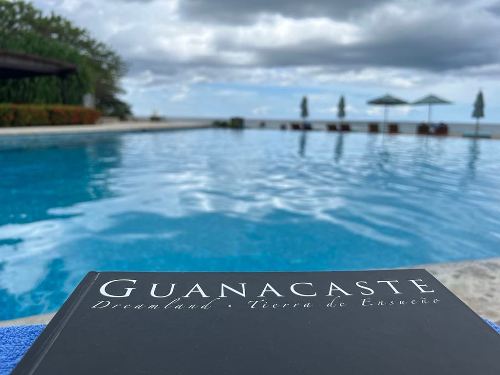 A book named "Gunacaste" near a beautiful, blue pool with the ocean in the back.