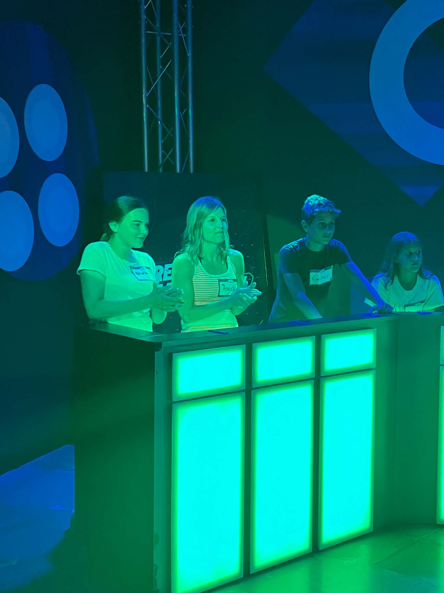 One woman, two teenagers and a preteen standing behind light up podiums where they are competing in a timed game.