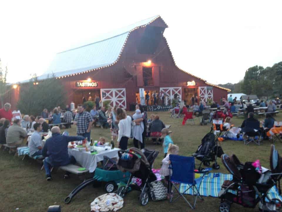 A barn with lights and people gathered all around having picnic dinners.