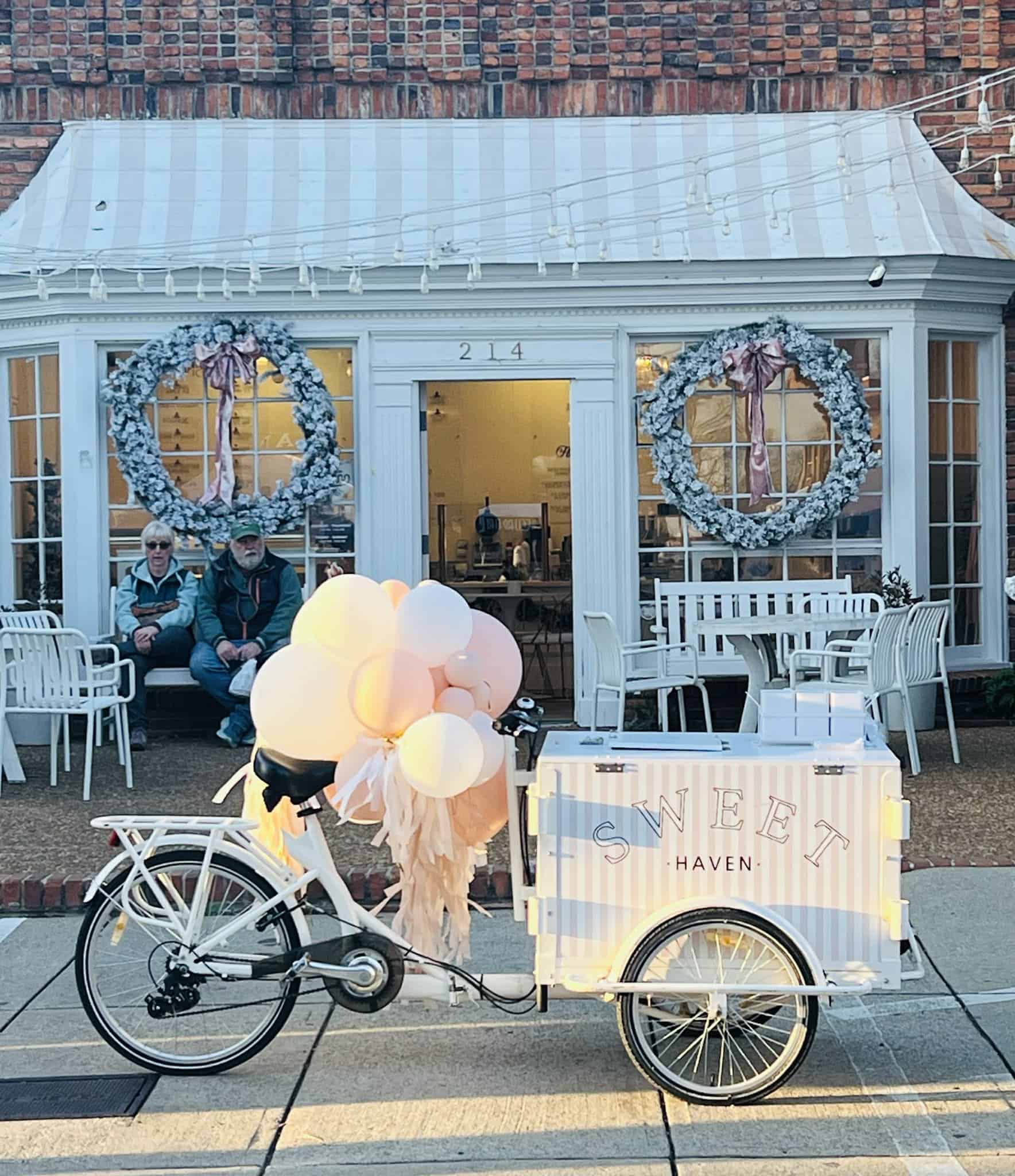 A bike with balloons and a pretty white cart in front of a beautiful ice cream shop.