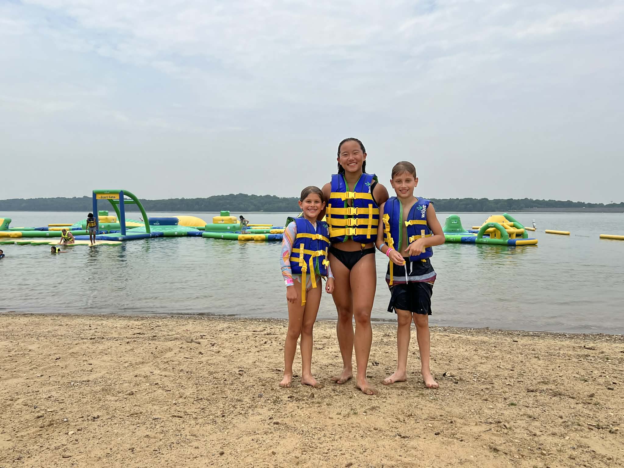 Two kids and a teenager with life vests on standing at the edge of the lake before they do the obstacle course.