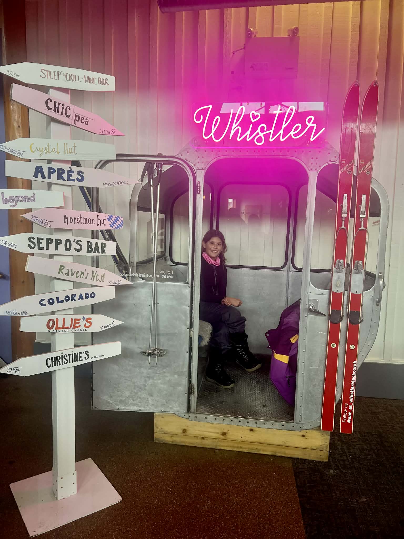 A little girl sits inside of an old gondola that has a neon pink "Whistler" sign above it. Next to her is a bunch of directional signs with names of other ski places.