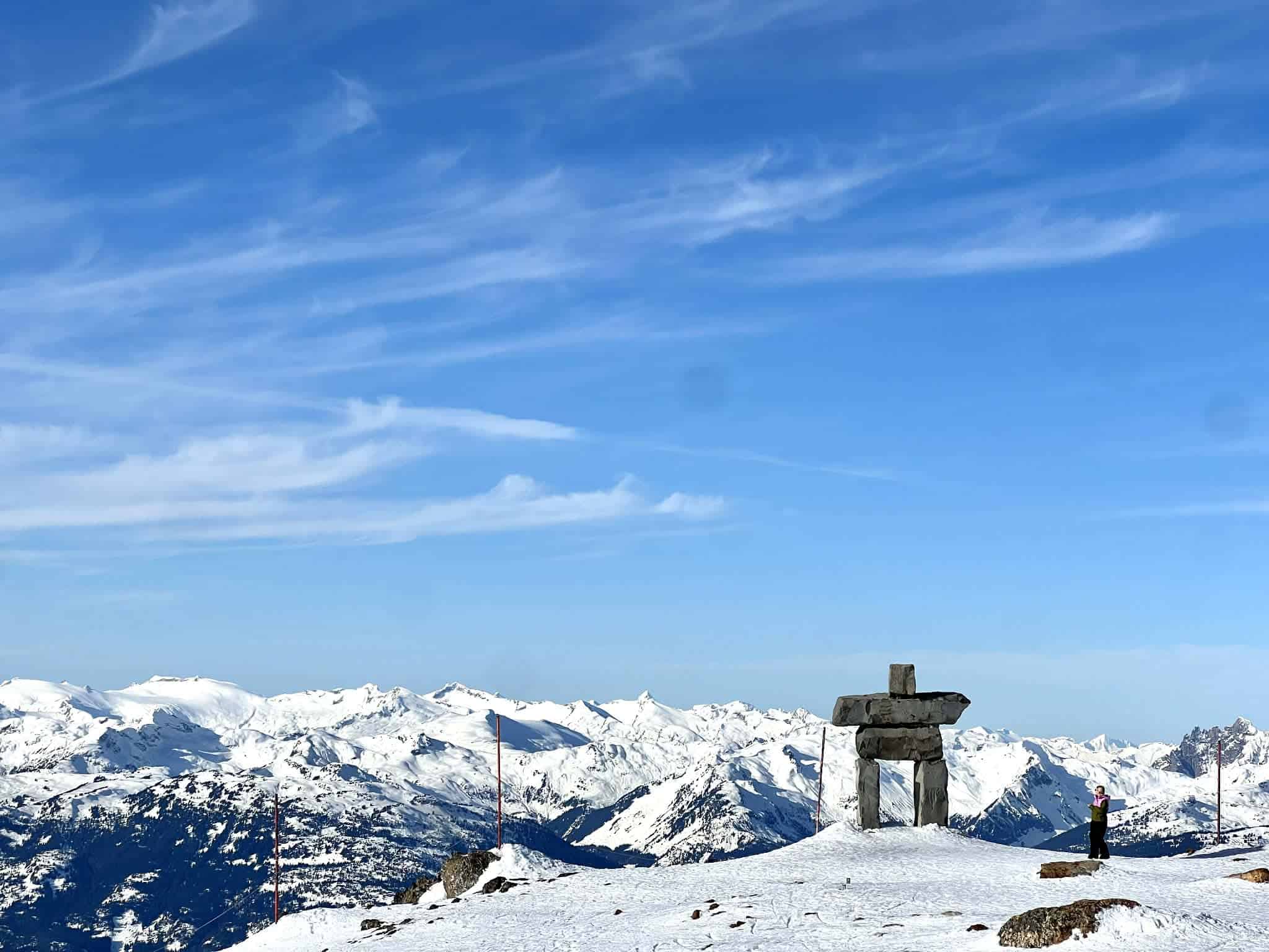 A stone statue shaped like a person is at the top of the mountain with a snow mountain range behind it.