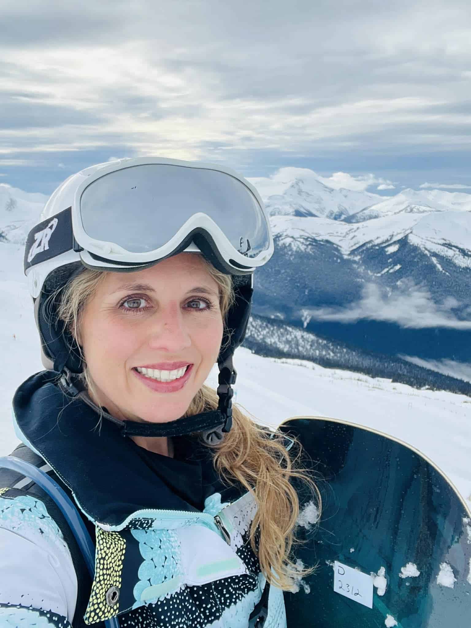 A woman stands with her snowboard and takes a picture of the mountains behind her.