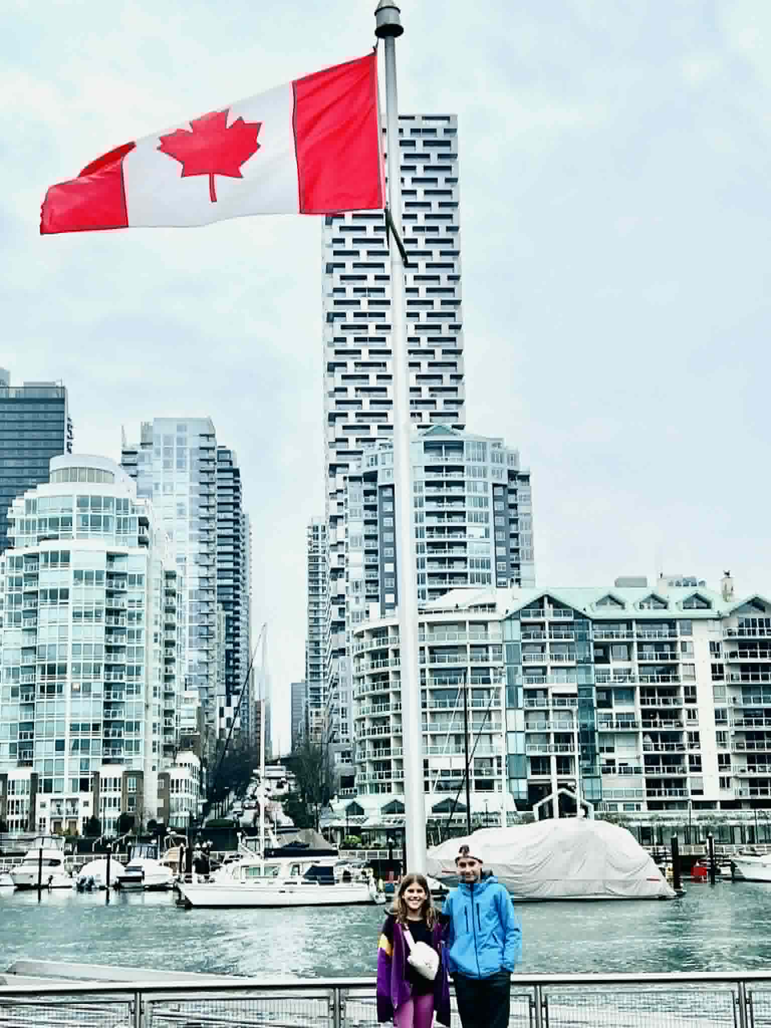 Two kids stand next to the Canadian flag with the Vancouver skyline behind them.
