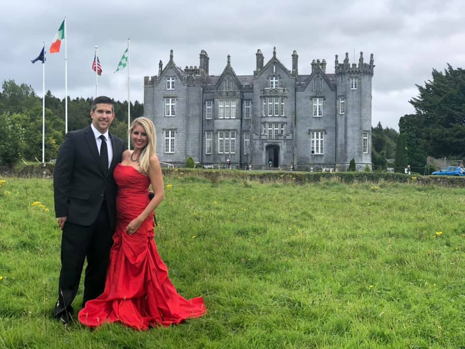 A man in a suit and a woman in a red dress standing in front of the Kinnity Castle in Ireland for the Sisterhood of the Castle Dresses.