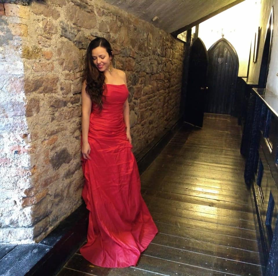 A woman standing against a stone wall at the Kinnitty Castle in Ireland. She is wearing the Sisterhood of the Castle Dresses red dress.