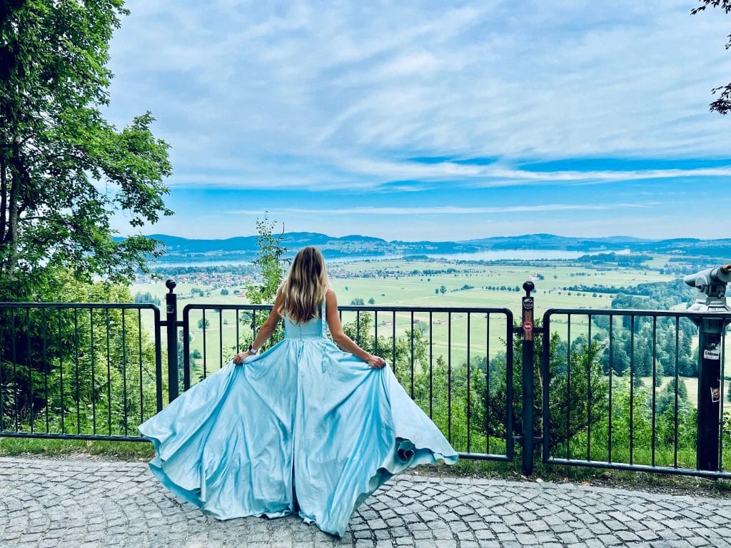 A girl in a blue dress at the Neuschwanstein Castle. The castle is not in the picture but she is overlooking the farmland in front of the castle. The blue dress looks like a princess dress.