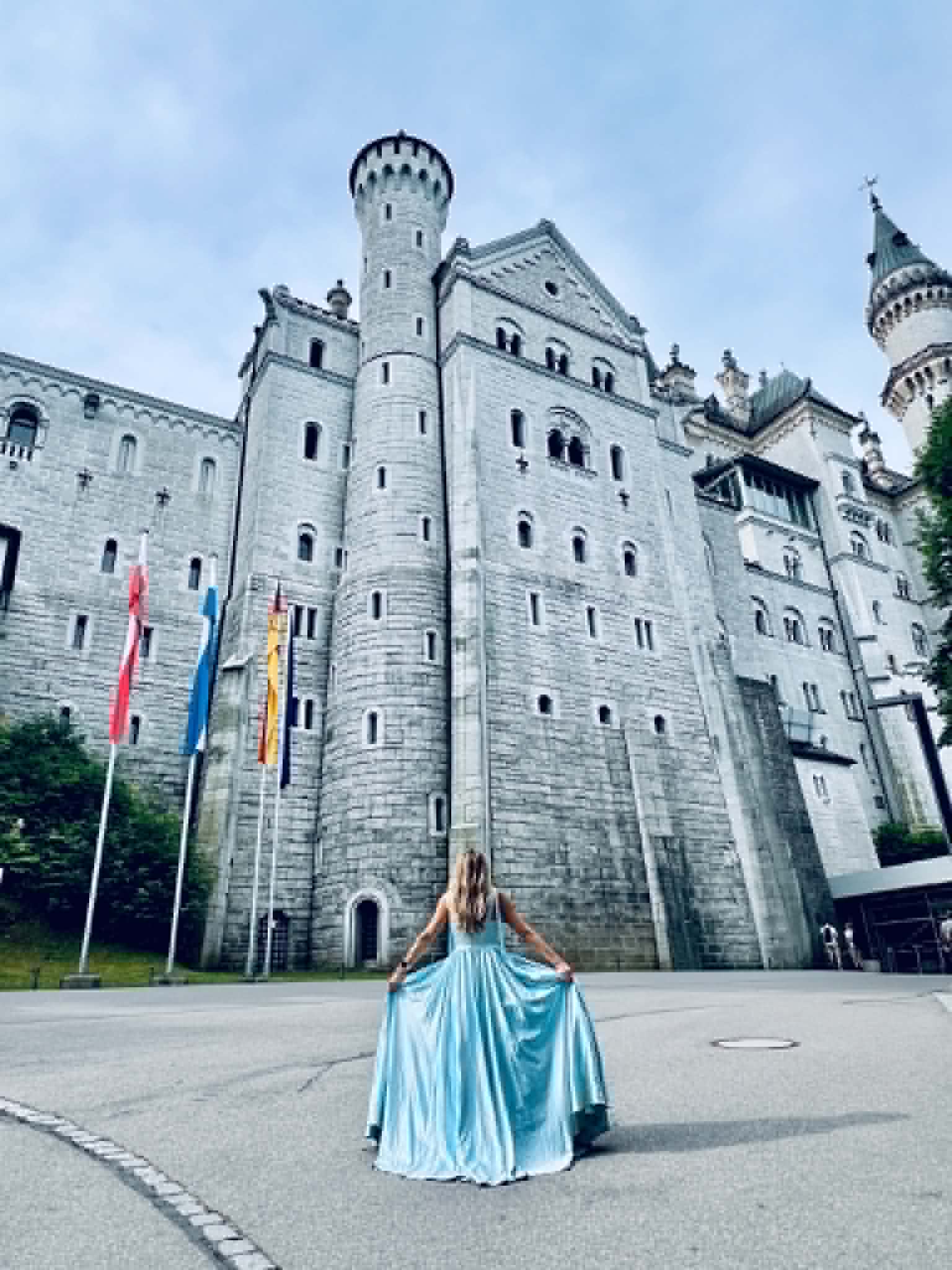 A woman in a big, blue princess style dress standing in front of the Neuschwanstein Castle in Germany. There are 3 flags also in the photo.