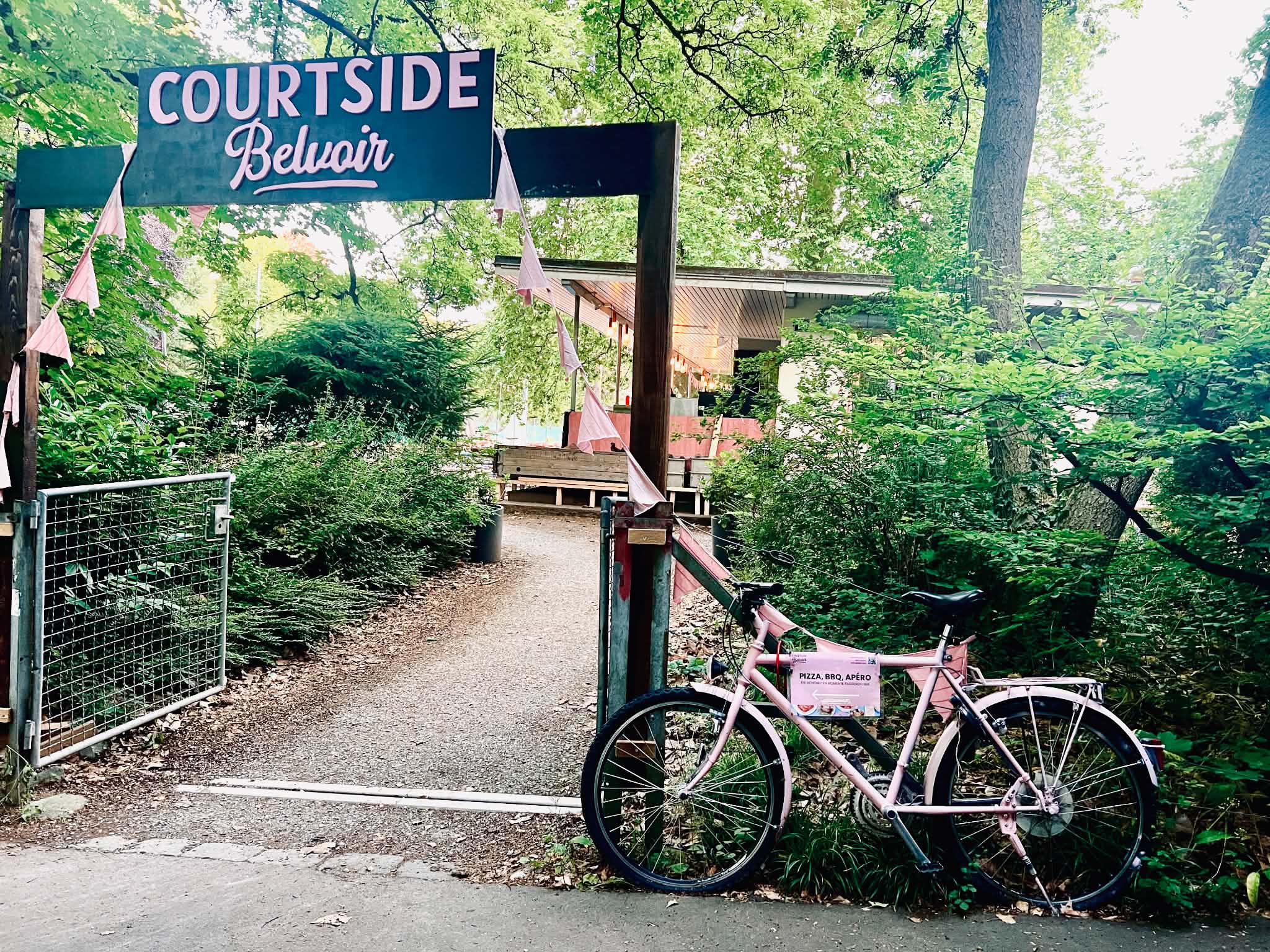 A picture of the entrance of Courtside Belvoir. It is a wooden sign with the letters in pink and a pink bicycle next to the entrance.