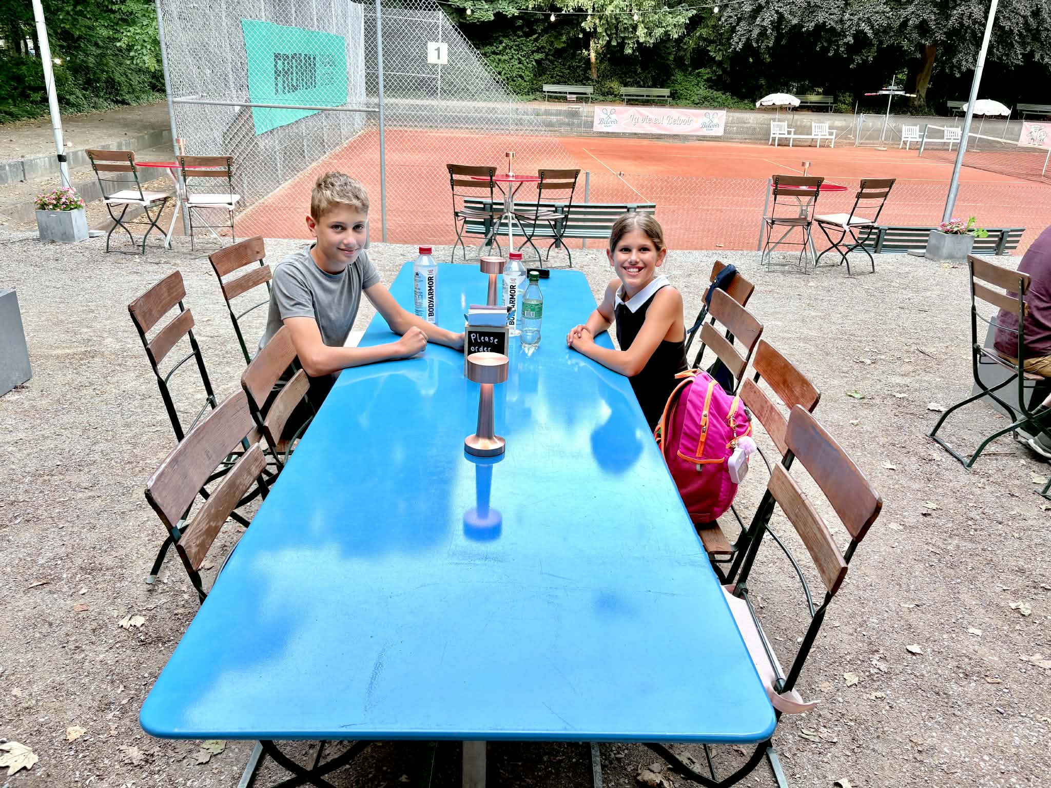 Two kids sitting at a blue long table outside waiting for their pizza. There is a tennis court in the background.