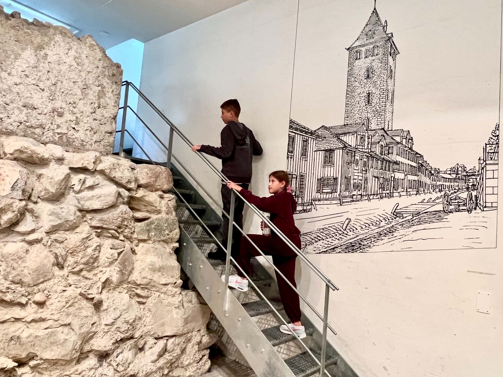 Two kids climbing stairs inside the Zurich Archeological Windows. There is really old stone to their left and a picture of what it used to be on the right.