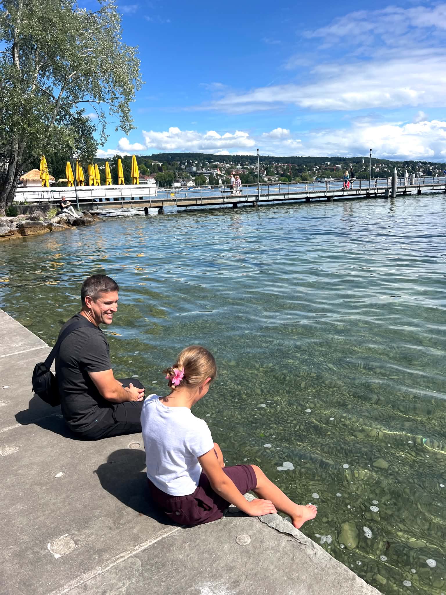 A dad sits with his daughter on the bank of Lake Zurich with their feet in the water.