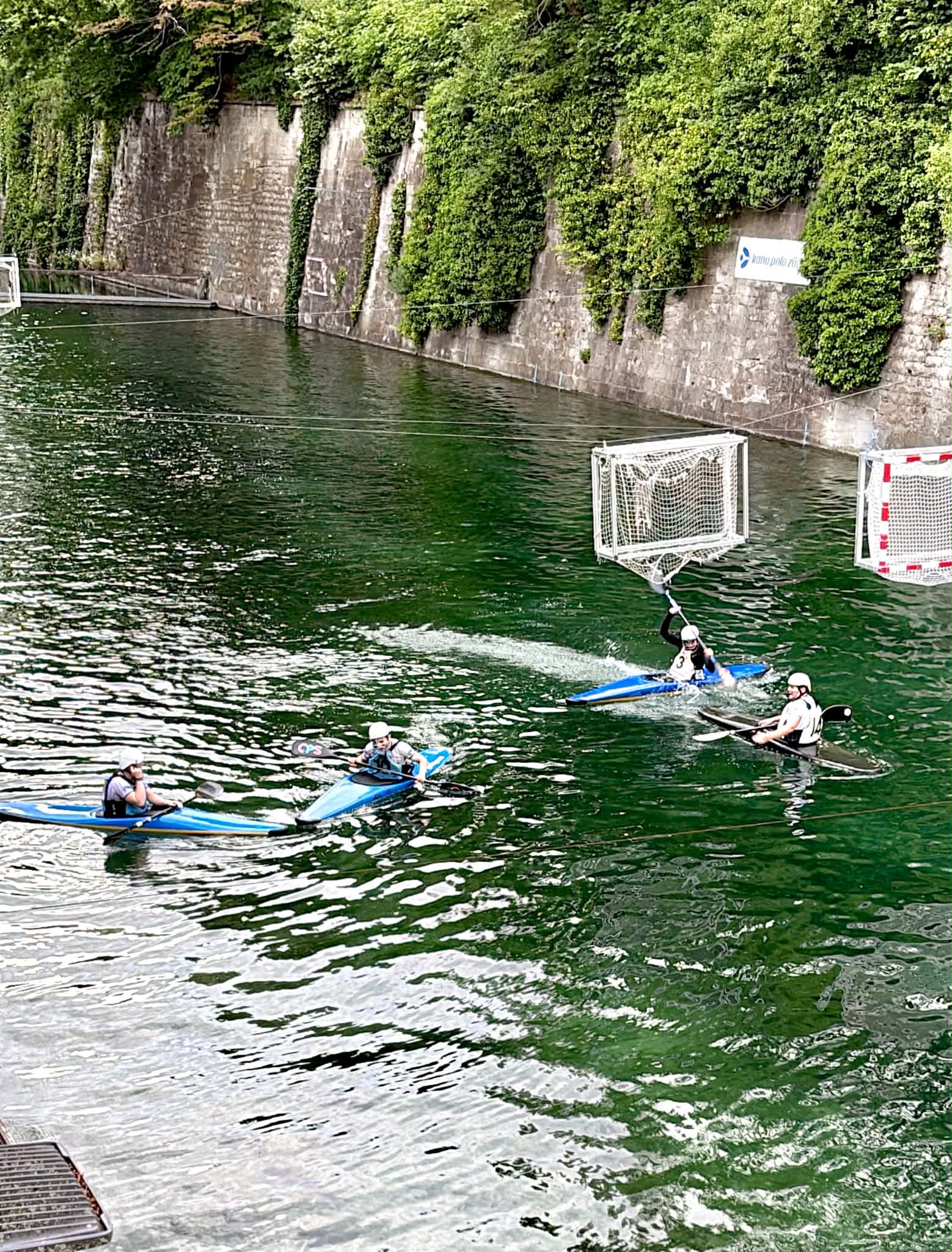 Four women playing "Canoe Polo" in a beautiful canal. There is greenery on the walls and nets hung above the canal.