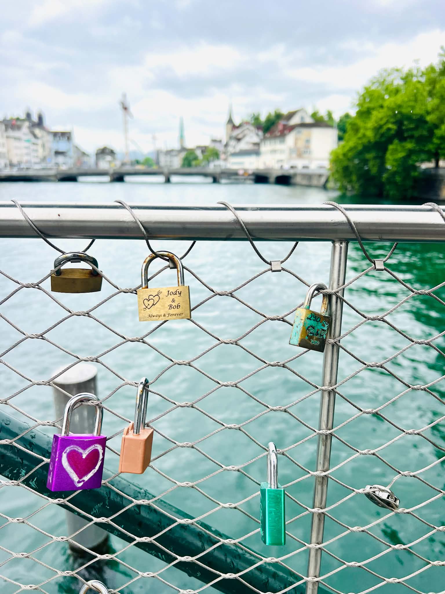 A picture of locks on a bridge with the water and another bridge and buildins behind it.