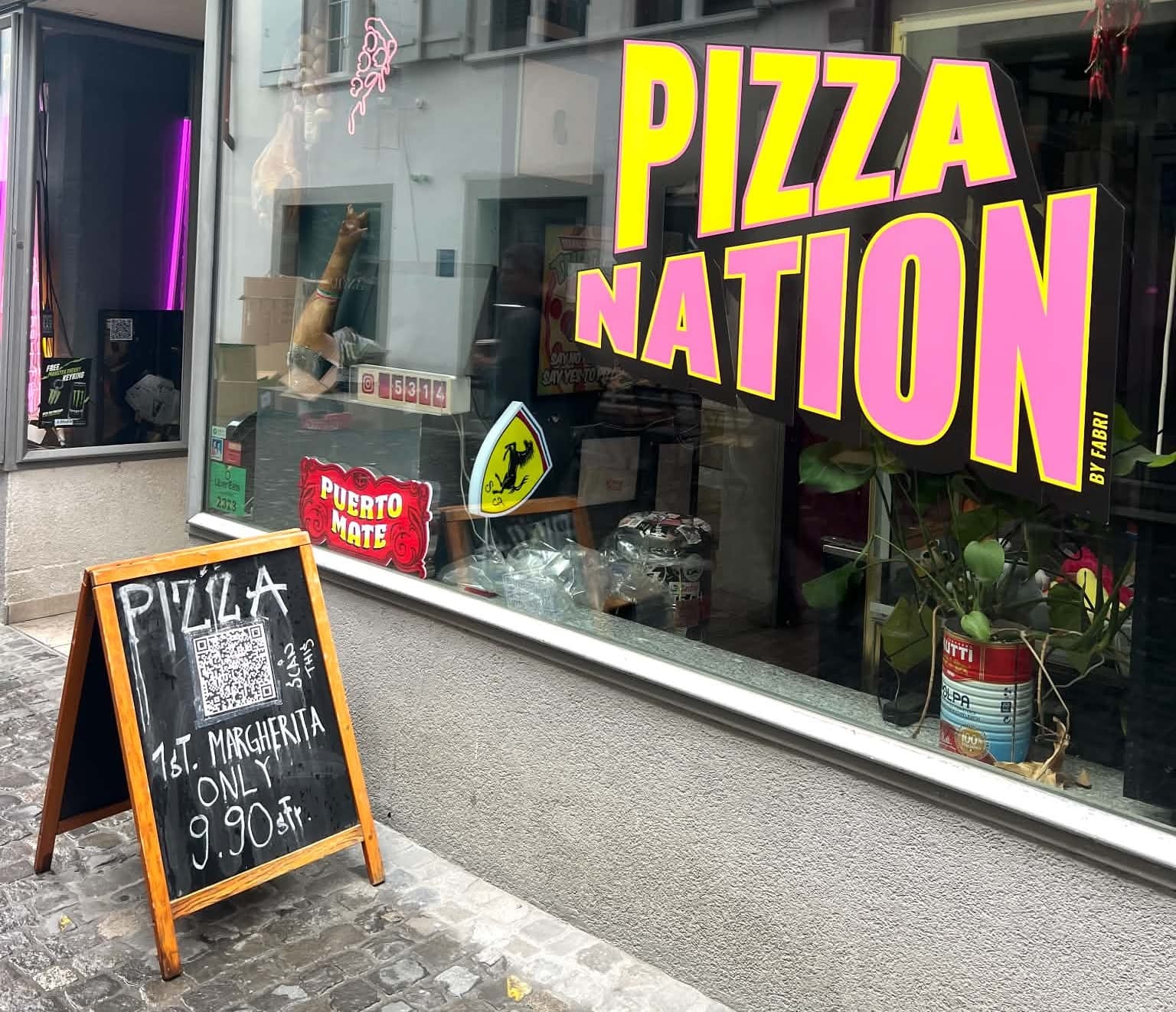 A picture of the outside of Pizza Nation. The words are really big and in pink and yellow. There is another sign giving the pizza special for the day.