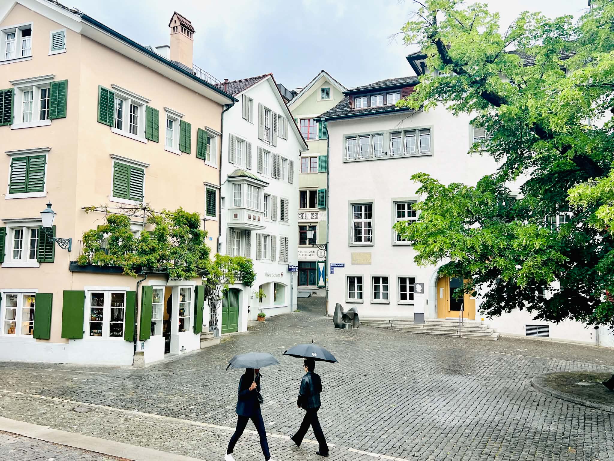 A really beautiful square in Zurich that has buildings and trees with two people walking with umbrellas in the rain.