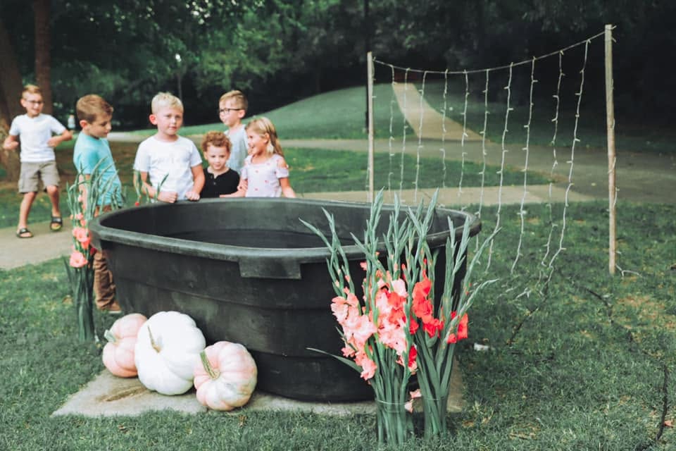 Kids look into the horse trough that has turned into a baptismal. It is decorated with white pumpkins, tall flowers and lights behind it in preparation for the baptisms.