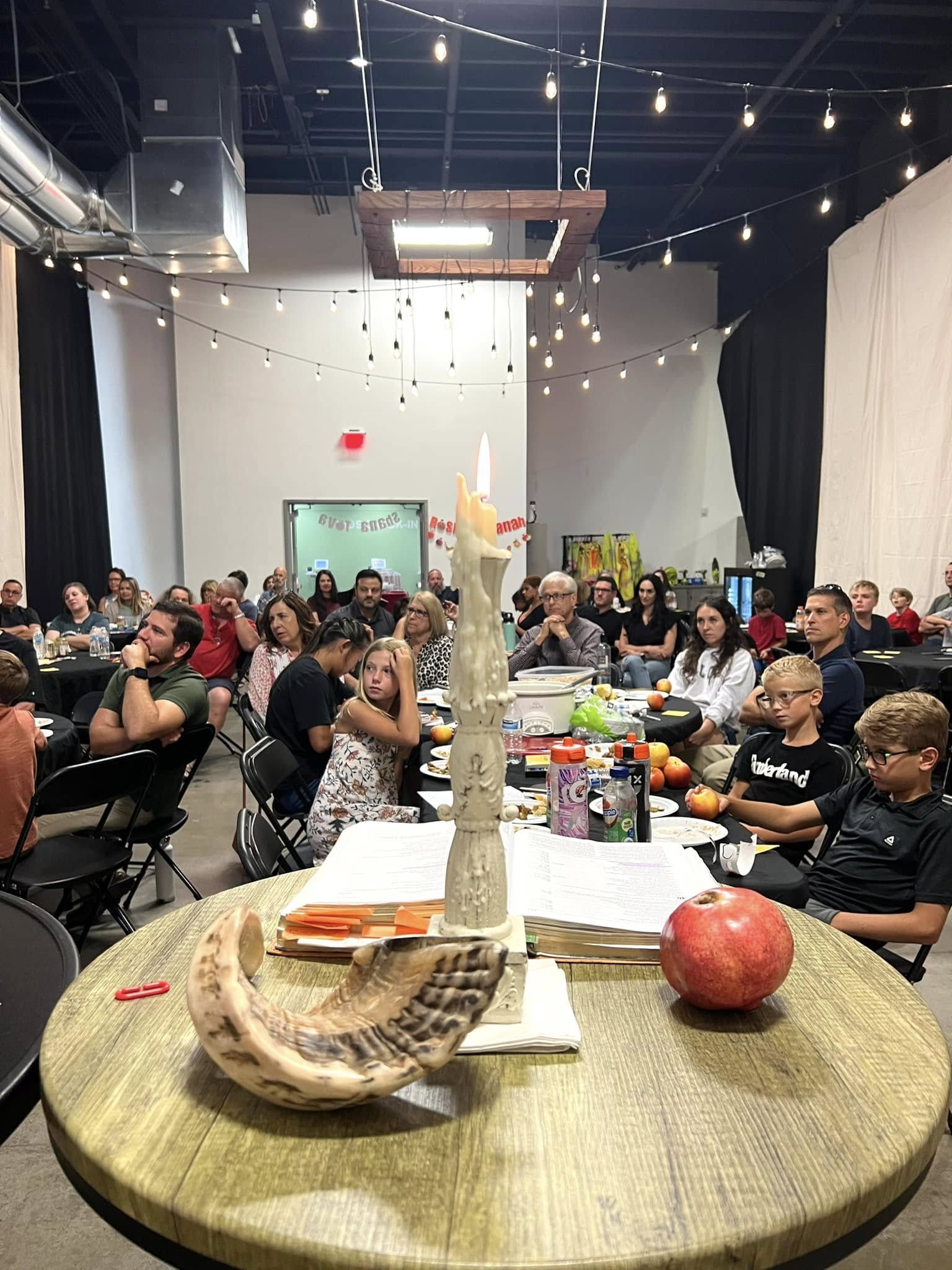 A room full of people sitting at round tables watching the presentation on why Feast of Trumpets is all about Jesus. There is a table in the front of the photo with a shofar, candle and Bible.