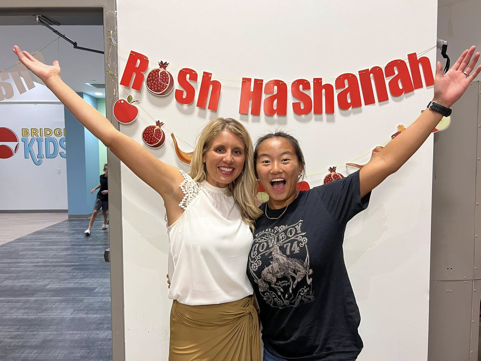 Two girls in front of a Rosh Hashanah sign with their arms spread wide.