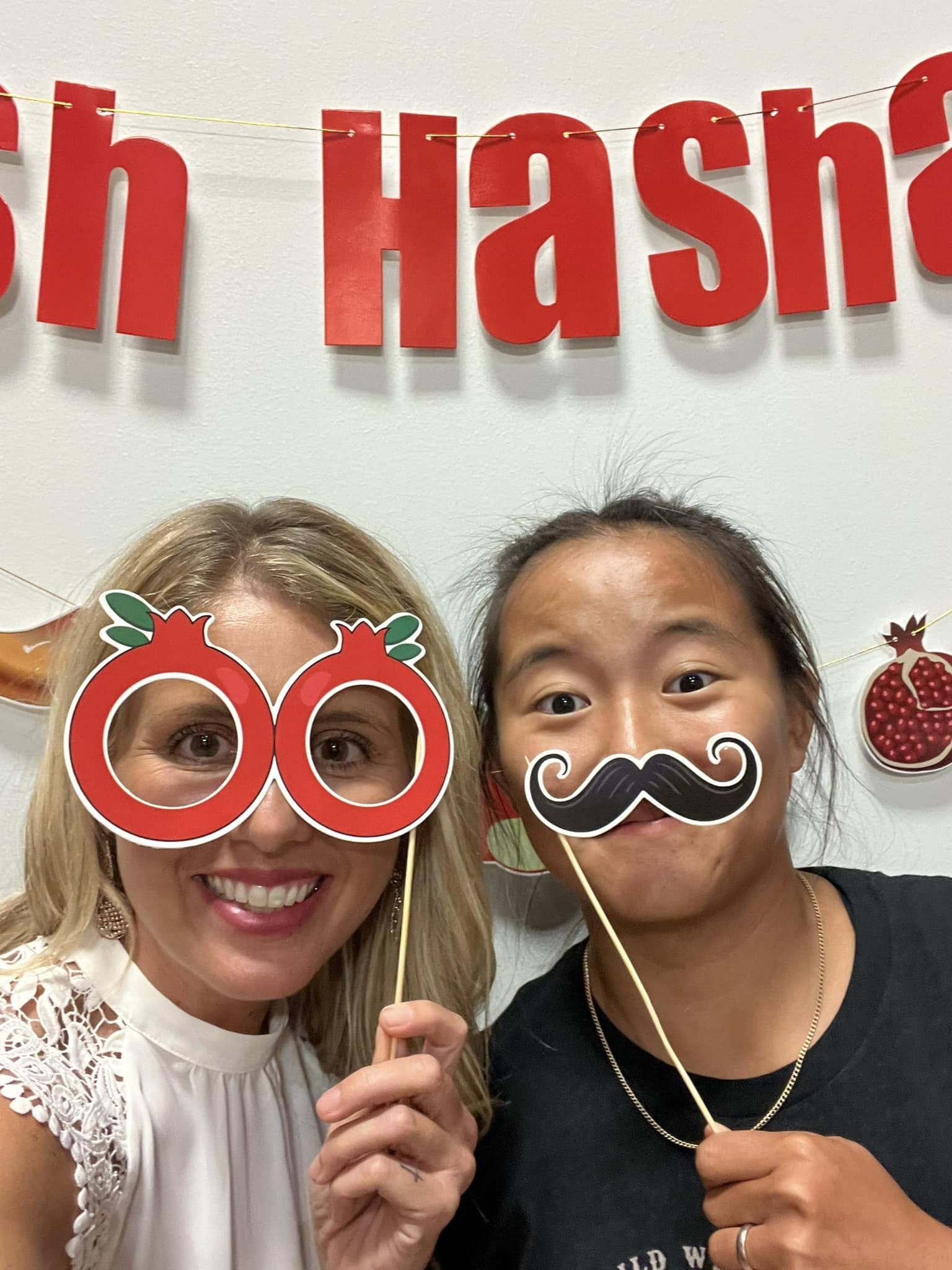 Two girls playing with the Rosh Hashanah decorations. One has pomegranate glasses and the other has a mustache photo prop.
